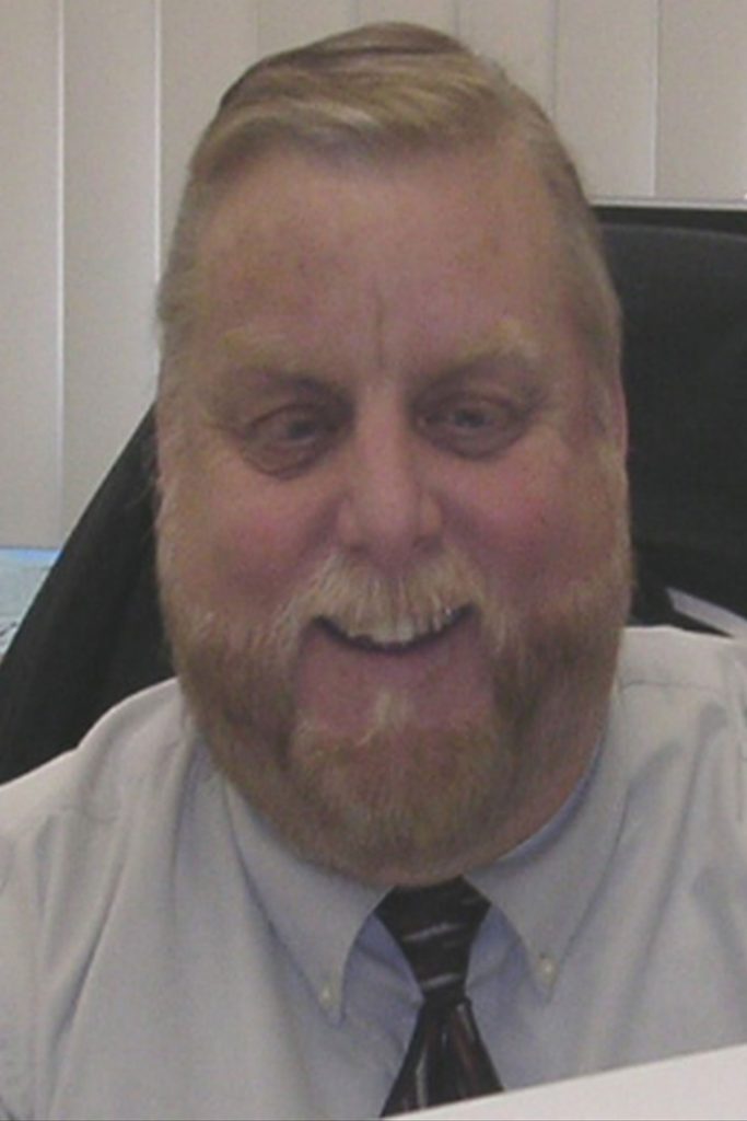 A man with a beard and mustache is sitting indoors, smiling. He is wearing a light-colored collared shirt with a tie, and there is a blurred background with vertical blinds behind him.