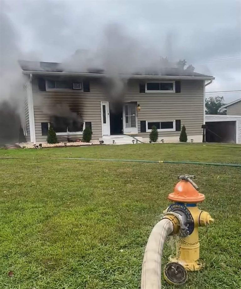 A split-level house with gray siding is emitting thick black smoke from an upper window and the front door. A fire hose is connected to a yellow fire hydrant on the lawn in front of the house. No visible flames, but the smoke suggests an active fire inside.