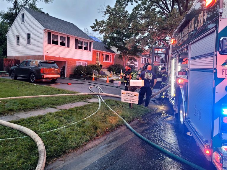 Firefighters respond to a house fire in a residential neighborhood. A fire truck with lights on is parked on the street, and hoses are laid out. A charred vehicle is in the driveway. Cones and a cleanup sign are visible on the lawn. It appears to be early evening.