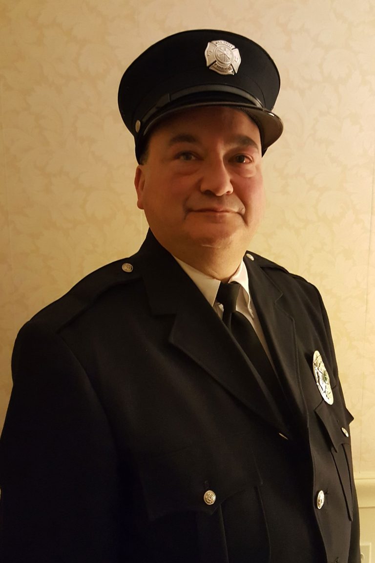 A person wearing a formal dark uniform and a peaked cap with a badge stands against a beige, patterned background. The uniform has insignias and buttons, suggesting an official capacity such as a firefighter or law enforcement officer.