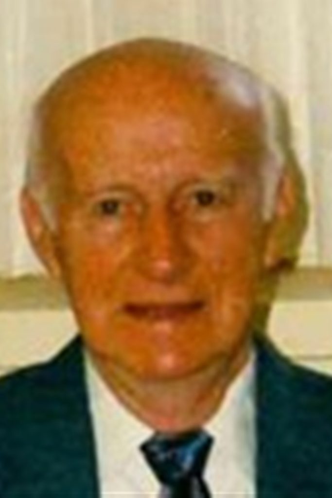 An elderly man with a bald head wearing a suit, white shirt, and dark tie, smiles slightly at the camera. The background appears to be light-colored curtains or drapes.