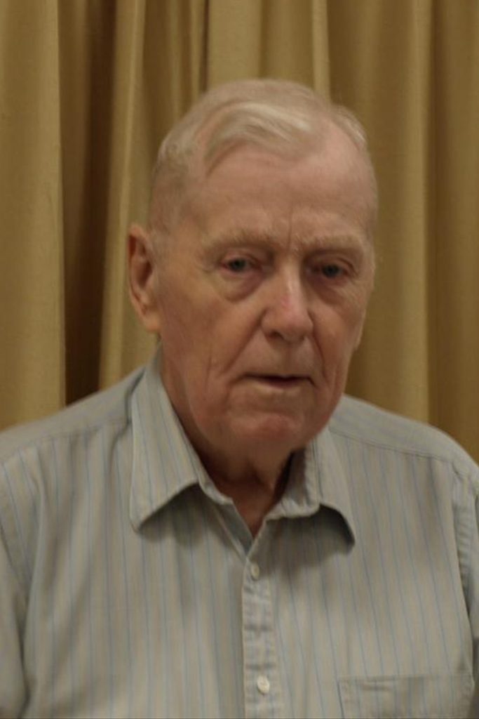 An older man with light hair is wearing a light blue, vertically striped shirt and standing against a plain beige curtain backdrop. His facial expression appears neutral, and he is looking slightly off-camera.
