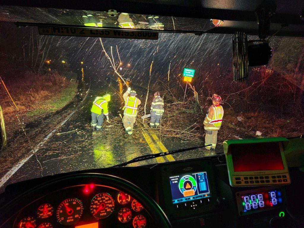 A nighttime scene viewed from inside a fire truck shows firefighters in reflective gear clearing downed tree branches from a wet road. The truck's dashboard, with illuminated controls and gauges, is visible in the foreground. Rain-splattered windshield adds depth.