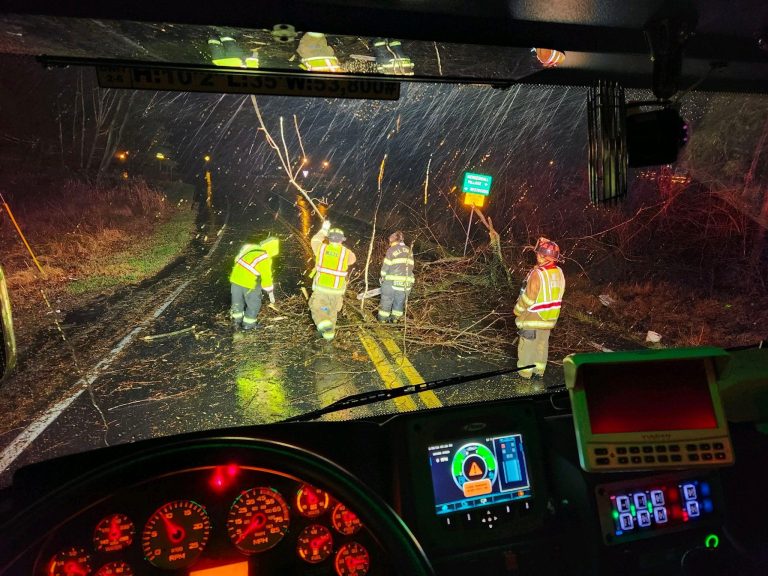 A nighttime scene viewed from inside a fire truck shows firefighters in reflective gear clearing downed tree branches from a wet road. The truck's dashboard, with illuminated controls and gauges, is visible in the foreground. Rain-splattered windshield adds depth.