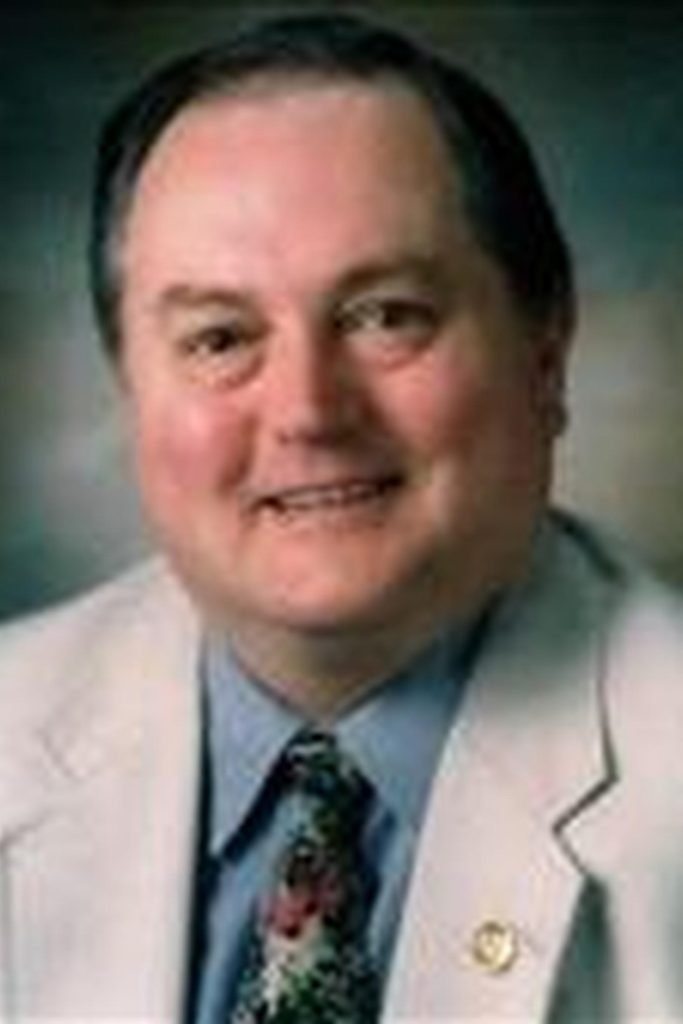 A middle-aged man with a friendly smile is wearing a light-colored suit jacket, a blue dress shirt, and a patterned tie. The background is blurry and neutral. He has short dark hair, and a lapel pin is visible on the left side of his jacket.