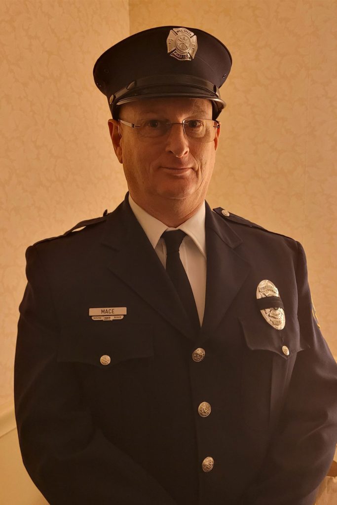 A person wearing a dark blue police or security uniform with a peaked cap stands indoors. The uniform features badges, a name tag saying "MACE," and buttons. The person is posed facing forward against a beige, patterned background.