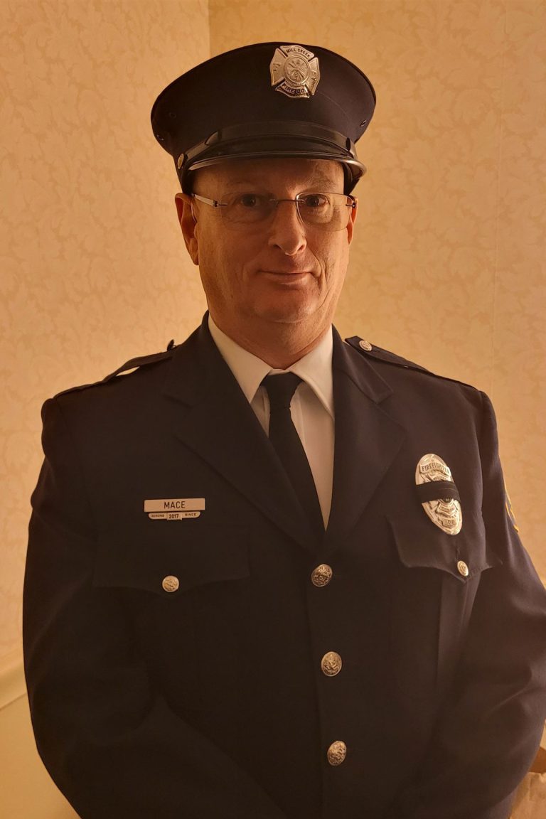A person wearing a dark blue police or security uniform with a peaked cap stands indoors. The uniform features badges, a name tag saying "MACE," and buttons. The person is posed facing forward against a beige, patterned background.