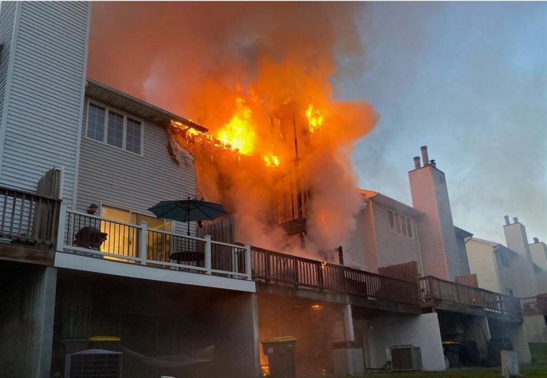 A house with white siding is engulfed in a fierce fire, with bright orange flames consuming the upper floor and thick smoke billowing into the sky. The wooden deck below is partially visible, with an umbrella and patio furniture still intact.