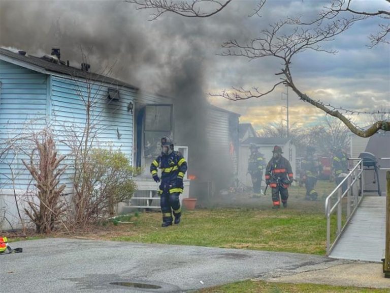 Firefighters in full gear exit a blue residential house with dark smoke billowing from its windows. The surrounding yard has some bushes and potted plants. The scene suggests an active firefighting effort under a cloudy sky.