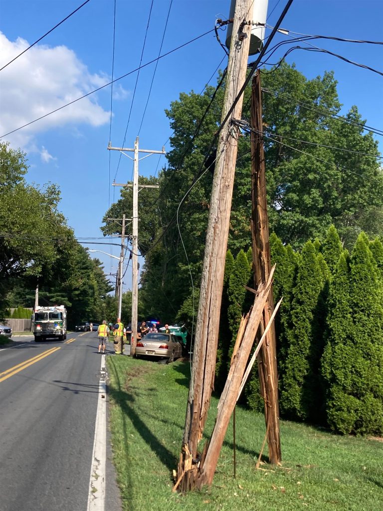 A street scene shows a large wooden utility pole broken and splintered at the base. Power lines dangle from the pole. Workers in reflective vests and emergency responders are on-site. A damaged car is partially visible behind bushes in a residential area.