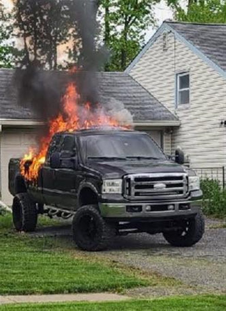 A black pickup truck engulfed in flames and emitting thick black smoke is parked on a gravel driveway in front of a garage. The surrounding area features a house with tan siding and a grassy lawn.