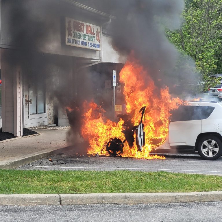 A white SUV is engulfed in flames and heavy black smoke in a parking lot near a storefront with a sign that reads "Great Wall Chinese Restaurant." The fire is intense, and the vehicle's interior and exterior are burning severely. A grassy area is in the foreground.