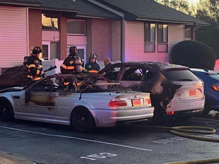 Firefighters in gear stand around two severely burnt cars in a residential parking lot, examining the scene. A hose lies on the ground near the cars. The adjacent building shows no visible damage. The setting is in front of apartment units numbered 153 and 154.