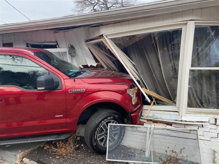 A red Ford F-150 pickup truck has crashed into the side of a white house, breaking through a large window and damaging the wall. Broken glass and debris are scattered around the scene.