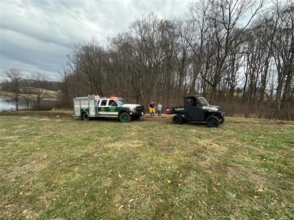 A grassy area near a forest with a lake in the background. A fire truck and an all-terrain vehicle are parked on the grass. Three people stand behind the fire truck. The weather is overcast and trees are bare, indicating winter or early spring.