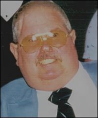 A man with short hair, wearing yellow-tinted glasses, a blue jacket, white shirt, and black tie, smiles at the camera against a dark background.