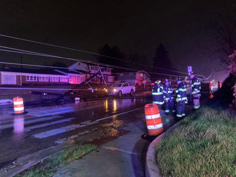 Nighttime scene of a car crash involving a white vehicle that hit a utility pole, causing it to lean over the street. Several firefighters in reflective gear are standing near the wreckage, with orange traffic barrels and cones on the wet road highlighting the area.