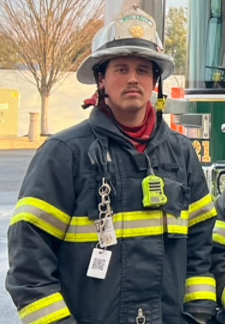 A firefighter stands in full gear, including a helmet and reflective striped jacket, with a communication device clipped to the front. A fire truck and bare trees in the background. The person has a serious expression.
