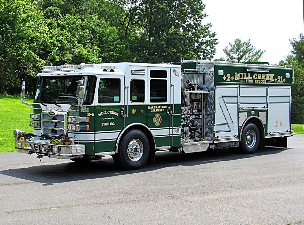 A green and white fire truck with the text "MILL CREEK FIRE CO." parked on a paved surface. The truck is equipped with various firefighting tools and equipment on its side and back. Lush green trees and grass are visible in the background under a clear sky.