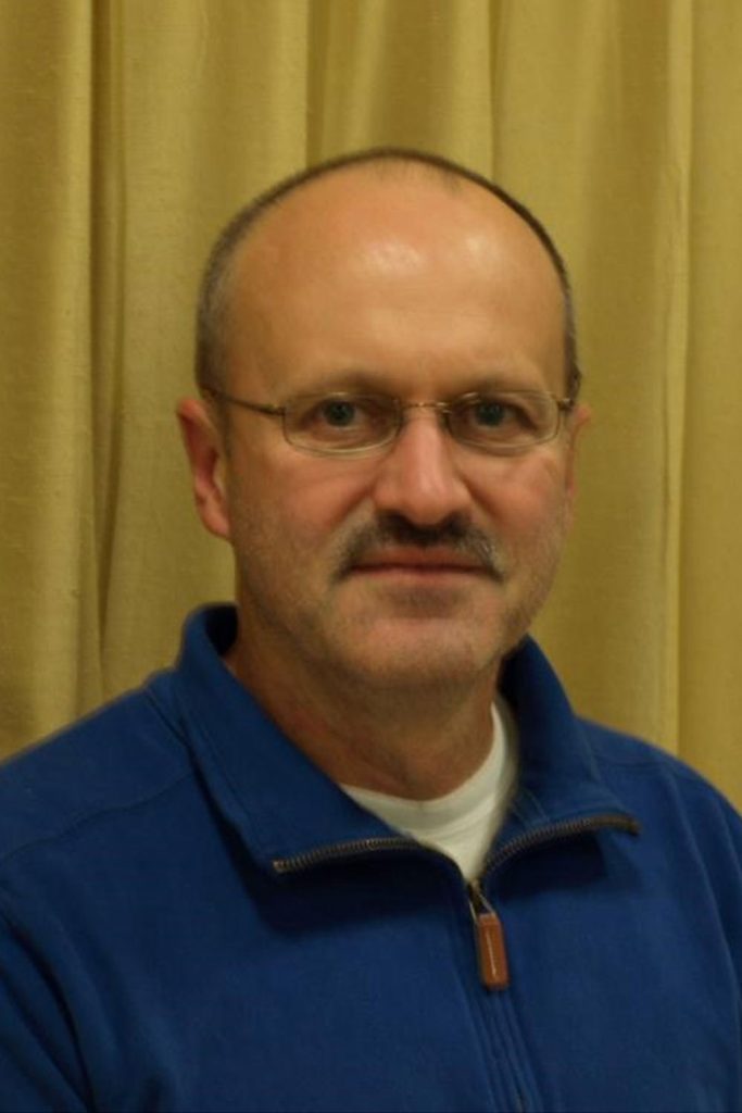 A man with a receding hairline, glasses, and a mustache is wearing a blue zip-up jacket over a white shirt. He is posing indoors in front of a beige curtain, looking straight at the camera with a neutral expression.