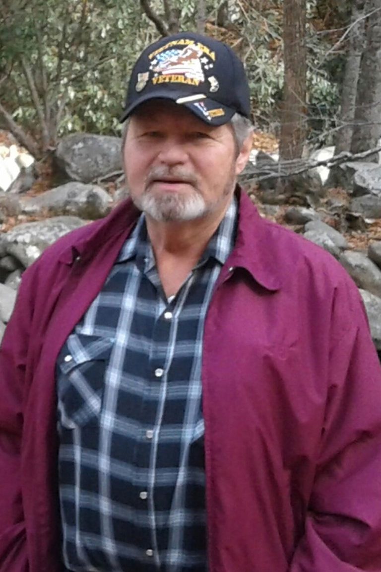 A man with gray hair and a beard is wearing a black hat that displays the word "Veteran" along with an emblem. He is dressed in a plaid shirt and a maroon jacket. The background consists of a forested area with rocks and trees.