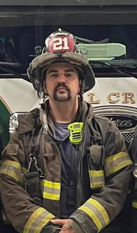 A firefighter wearing a helmet labeled "21," full gear, and a yellow radio stands in front of a vehicle with part of a logo visible behind him.
