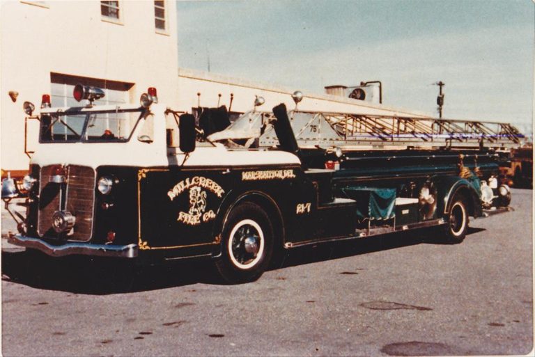 A vintage fire truck from Mill Creek Fire Company, painted in black and white, is parked outside a building. The side of the truck has a golden emblem that reads "Mill Creek Fire Co." and displays a large, extendable ladder on top.