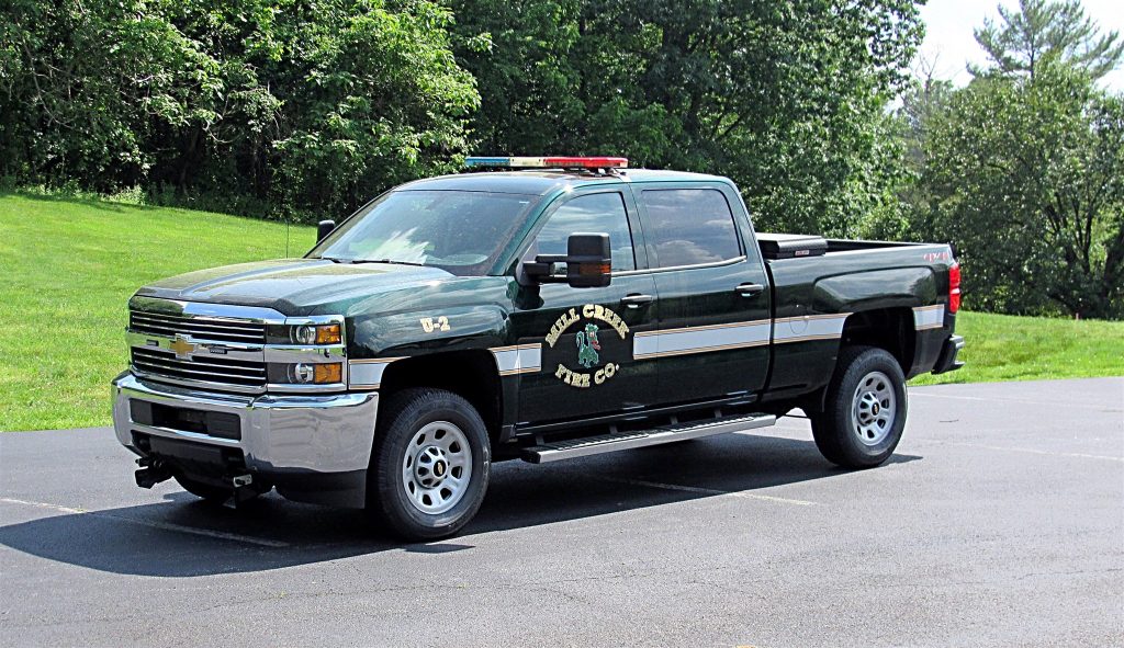 A green Chevrolet Silverado pickup truck with a "Royal Chevrolet Fire Co." logo on the door is parked on an asphalt surface surrounded by a lush, green, tree-filled landscape. The vehicle is equipped with emergency lights on the roof.