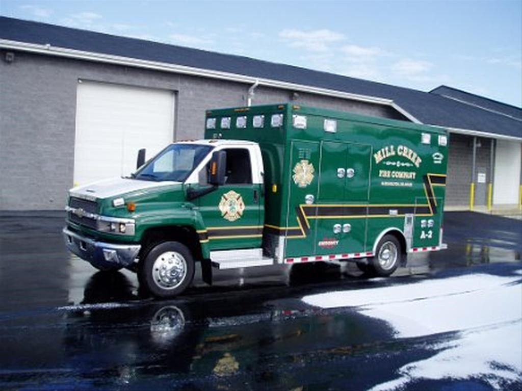 A green and white Mill Creek Fire Company ambulance is parked outside a brick building with a white garage door. The vehicle has gold and white markings and numbers, and the ground is wet with patches of foam or soap. The sky is clear.