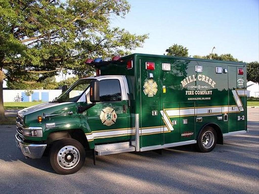 A large green ambulance from Mill Creek Fire Company, labeled "B-2", is parked on a paved area. The vehicle has emergency lights on top and graphic decals on the side, under a partly cloudy sky and nearby trees.