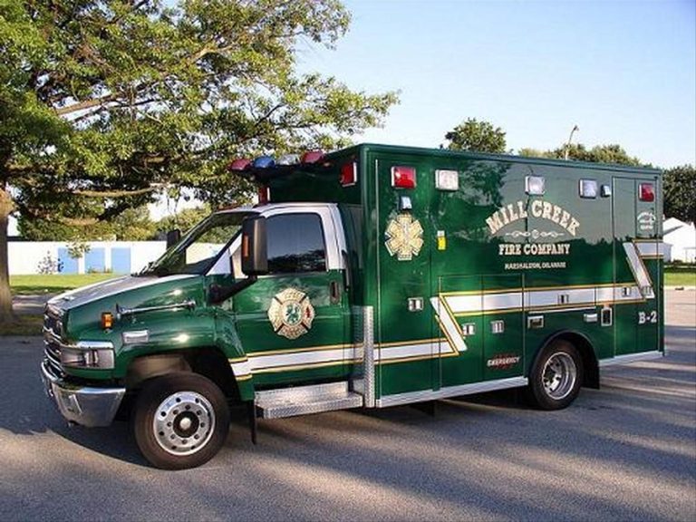 A large green ambulance from Mill Creek Fire Company, labeled "B-2", is parked on a paved area. The vehicle has emergency lights on top and graphic decals on the side, under a partly cloudy sky and nearby trees.