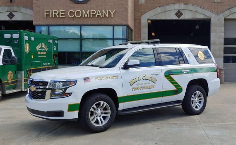 White SUV with green and yellow stripes parked in front of a fire company building. The vehicle is labeled "Duty Officer" and "Mill Creek Fire Company" with its logo on the doors. An emergency vehicle is partially visible in the background.