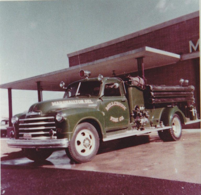 A vintage green fire truck with the text "Marshallton Del. Fire Co." on its side is parked in front of a building with a flat roof. The building has "M" visible on the wall, suggesting it could be an old fire station. The photo has an aged, 1950s look.