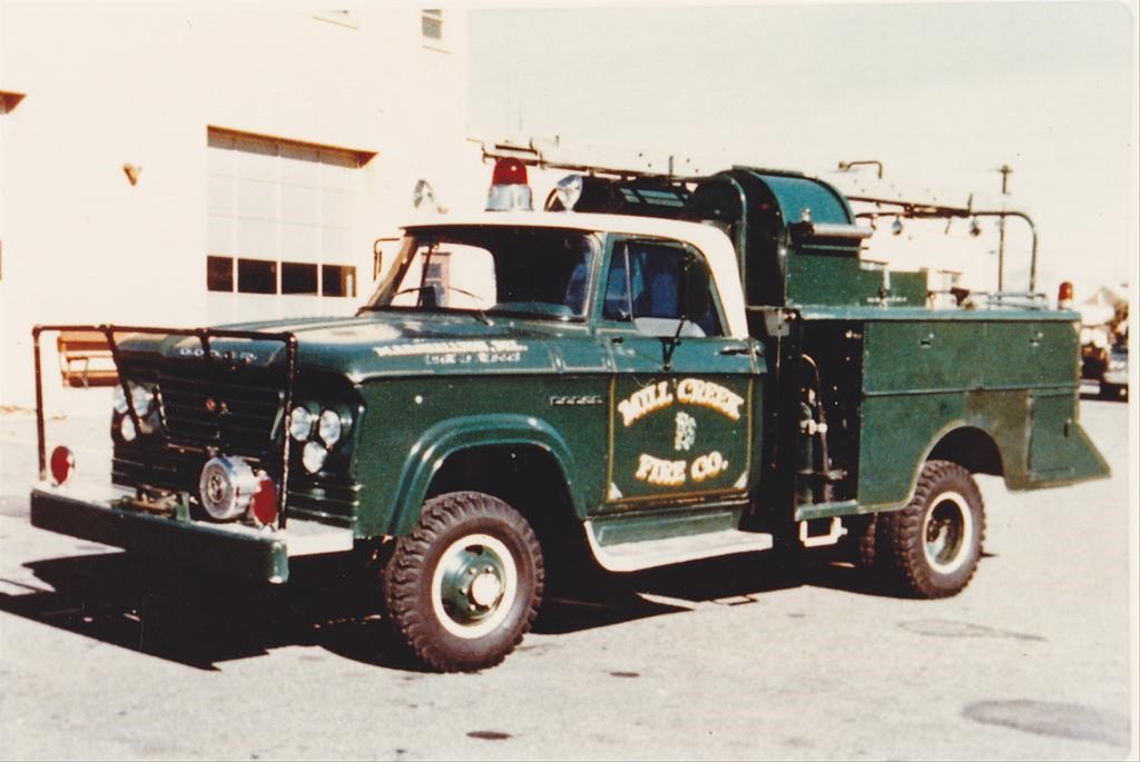 A vintage green fire truck from Mill Creek Fire Co. is parked outdoors. The truck features an old-fashioned design with equipment and storage compartments for firefighting, and a text logo on the side door. A fire station building is visible in the background.