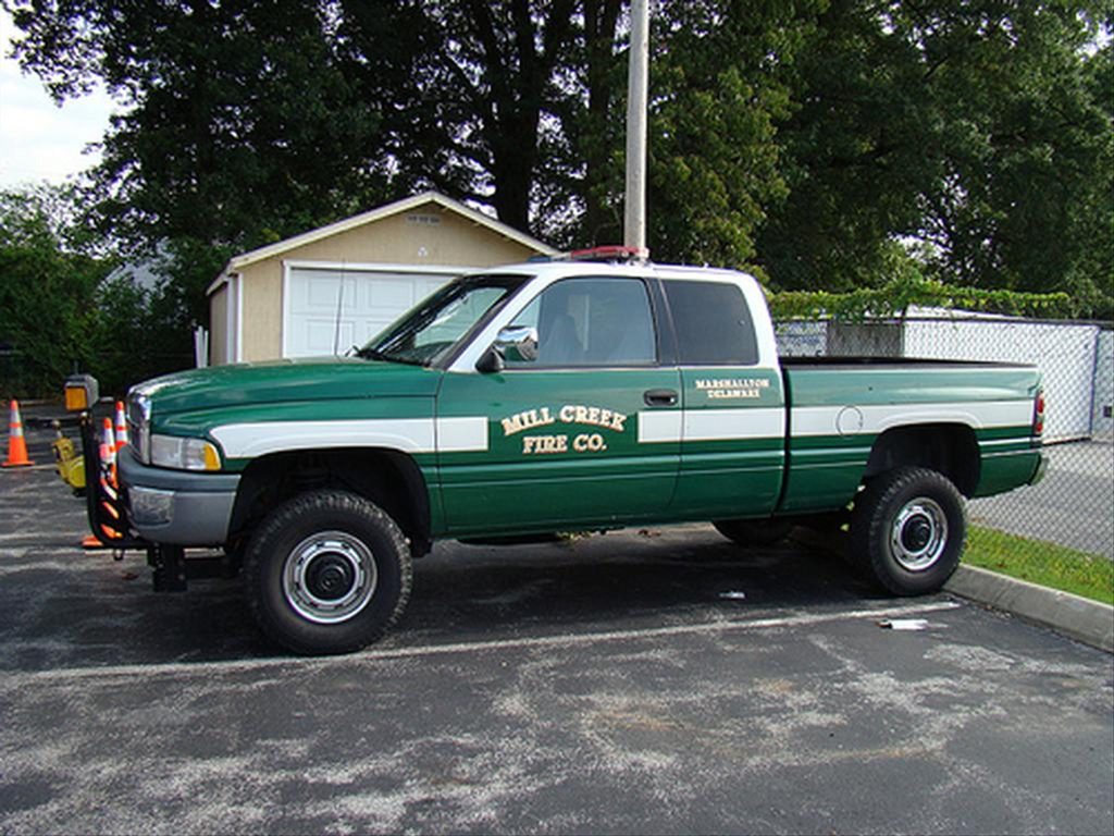 A green and white pickup truck with "Mill Creek Fire Co." and "Marshallton Delaware" written on the side is parked in a lot. The truck features emergency lights on top and is situated next to a small shed and some traffic cones, with trees in the background.