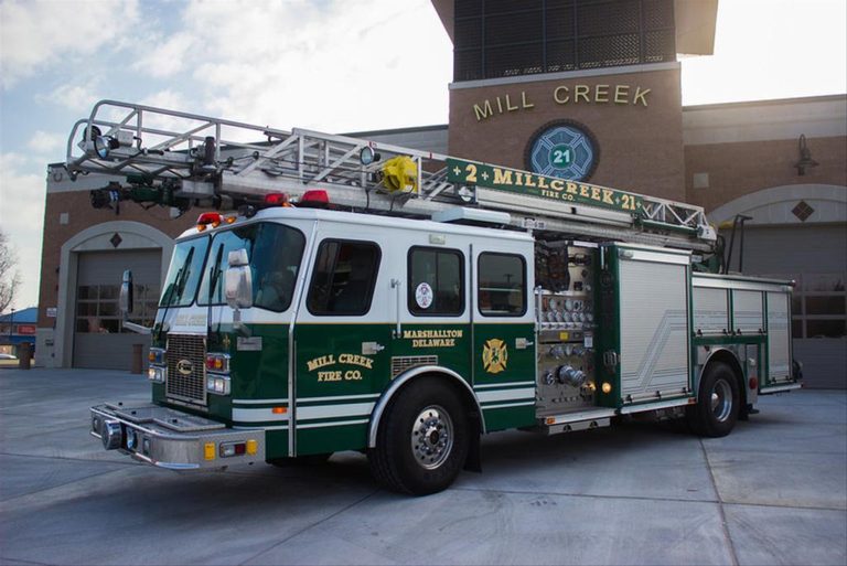 A green and white fire truck from the Mill Creek Fire Company is parked in front of a fire station with a sign that reads "Mill Creek" above the garage bay doors. The fire engine has an extended ladder mounted on top.