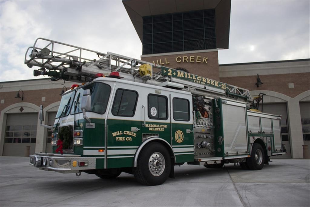 A green and white fire engine from Mill Creek Fire Co. is parked in front of a fire station. The fire truck is equipped with various tools and a ladder on top. The station's name is visible on the building's exterior. The sky is cloudy.