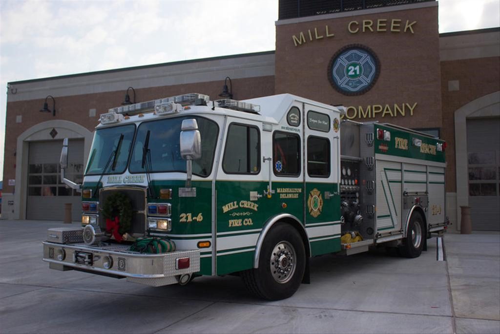 A green and white fire truck is parked on a concrete surface in front of the Mill Creek Fire Company building. The truck displays "Mill Creek Fire Co." and "21-6" branding. The building has an arched entrance with the number 21 above it.