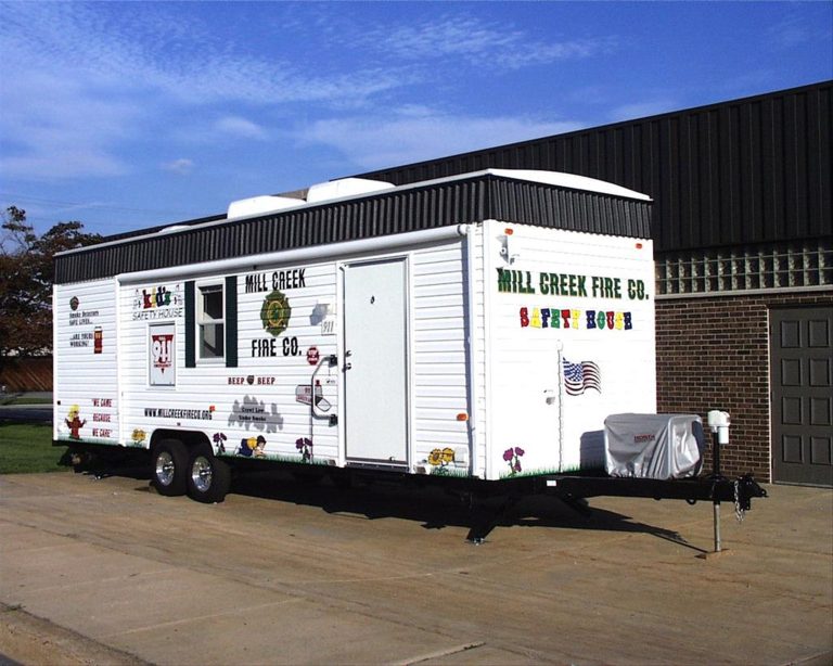 A white mobile trailer labeled "Mill Creek Fire Co. Safety House" with various decorations and logos is parked on a concrete surface near a large brick building. An American flag is attached to the trailer.