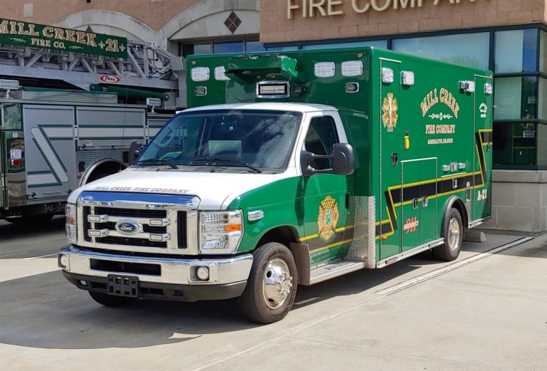 A green and white ambulance with the label "Mill Creek Fire Company" on the side door is parked outside a fire station. The vehicle is adorned with yellow and black stripes and a crest near the front. The fire station has signage and a garage with emergency vehicles inside.