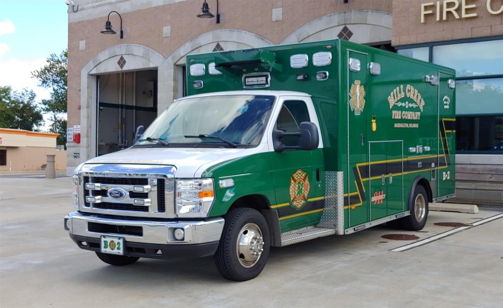 A green ambulance with "Hill Country Fire Chief" and "EMS" markings parked in front of a fire station. The vehicle features emergency lights, equipment compartments, and a custom license plate with "B02". The fire station facade includes beige brickwork and black lamps.