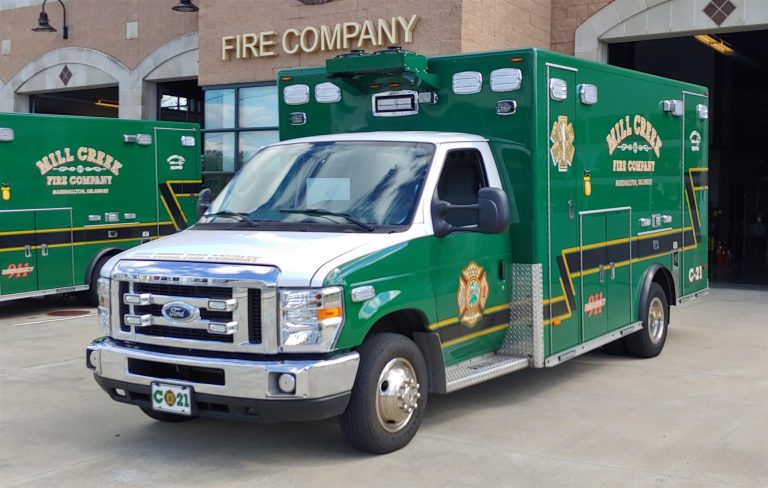 A green and white ambulance with "Mill Creek Fire Company" written on the side is parked in front of a fire station. The vehicle has emergency lights on top and the station building is visible in the background with a sign reading "FIRE COMPANY.