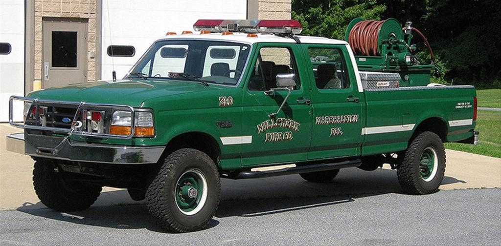 A green fire department pickup truck with "619" on the side and equipped with firefighting gear is parked outside a garage. The truck has a front push bar, emergency lights on the roof, and a hose reel mounted in the bed.