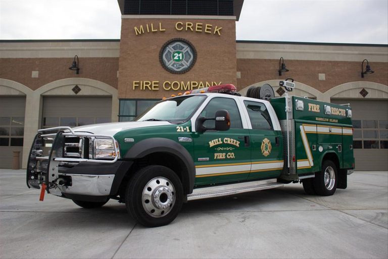 A green and white fire rescue truck with the label "Mill Creek Fire Co." is parked in front of the Mill Creek Fire Company building. The truck features emergency lights and equipment on the sides. The building has a large yellow emblem and multiple garage doors.