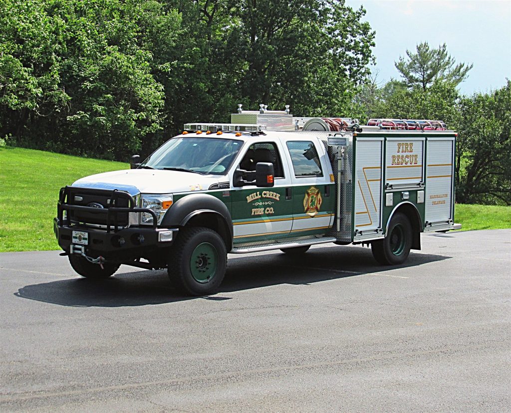 A white and green fire rescue truck from Mill Creek Fire Company is parked on a paved surface with lush green trees and grass in the background. The truck has emergency lights on top and various equipment stored on the sides.
