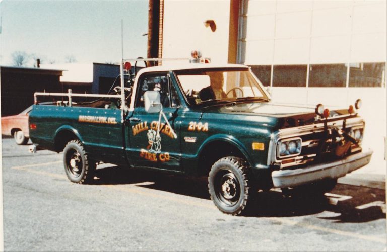 An old fire department pickup truck is parked in front of a building. The green truck, labeled "Mill Creek Fire Co." on the door, appears vintage with firefighting equipment on the back. The truck's markings indicate it is from Hockessin, Delaware, and have the number "2704".