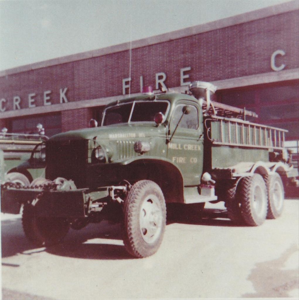 Vintage photo of a green military-style fire truck parked in front of a building with the words "Mill Creek Fire Co" partially visible on both the truck and the building. The truck is equipped with a ladder and has six large wheels.