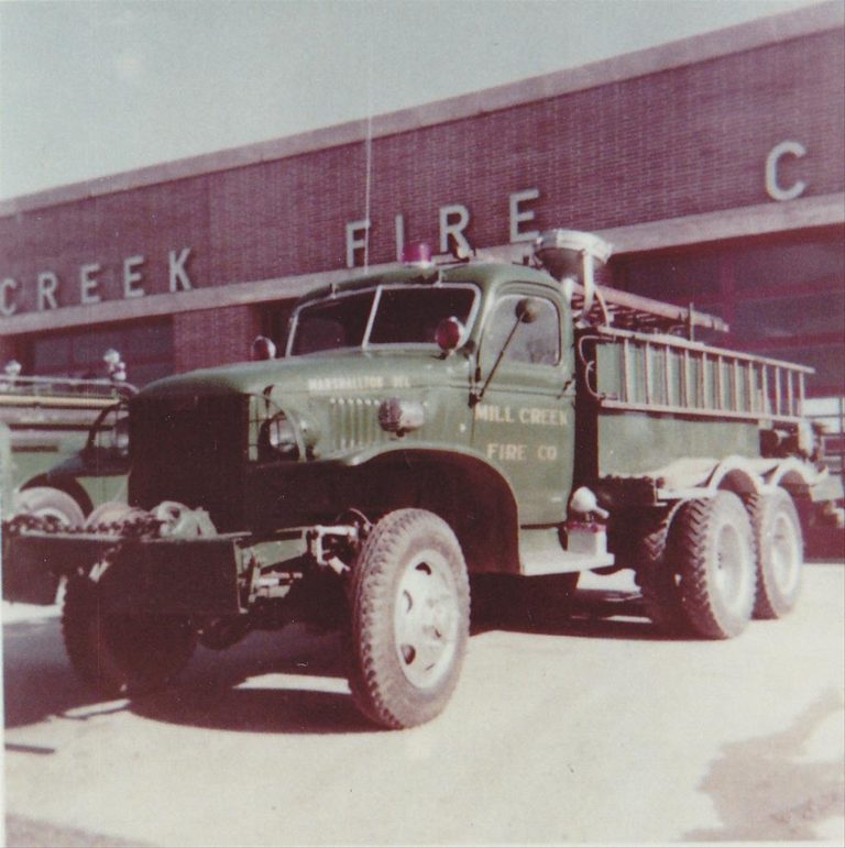 Vintage photo of a green military-style fire truck parked in front of a building with the words "Mill Creek Fire Co" partially visible on both the truck and the building. The truck is equipped with a ladder and has six large wheels.
