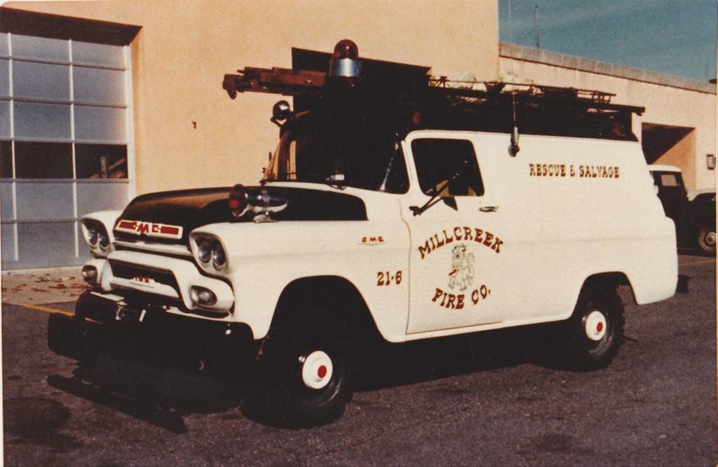 A vintage GMC fire and rescue vehicle from Millcreek Fire Co. is parked outside a building. The truck is white with "MILL CREEK FIRE CO." and "RESCUE & SALVAGE" written on the side. It has a red light on top and a black grill guard.