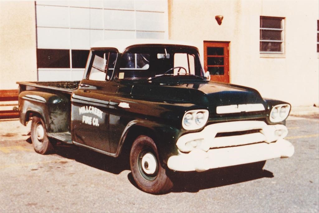 Vintage dark green pickup truck with "WILL-GREEN PAYNE CO" written on the door, parked in front of a beige building with large windows and a red door. The truck has a classic design with white trim on the front bumper and large, round headlights.